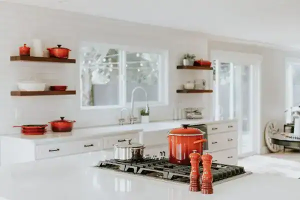 Modern white kitchen with red cookware and natural light from large windows.