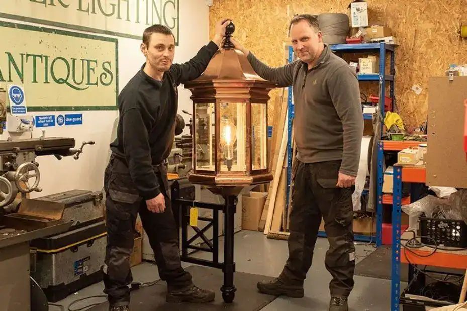 Two men holding a vintage lantern-style light fixture in workshop.