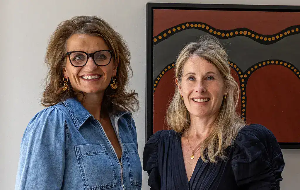 Two women smiling and standing in front of a colourful abstract artwork at Fritz Fryer Lighting UK.