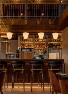 4 white glass slender silhouette pendants with sleek curves starting with a wide top and narrower bottom, suspended from 4 wall brackets above a modern wooden curved bar.