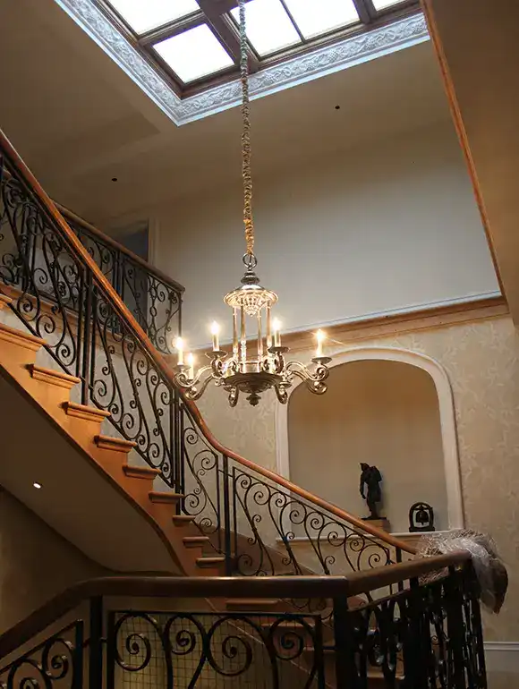 Chandelier lighting fixture hanging above a curved staircase with ornate iron railing and a skylight.